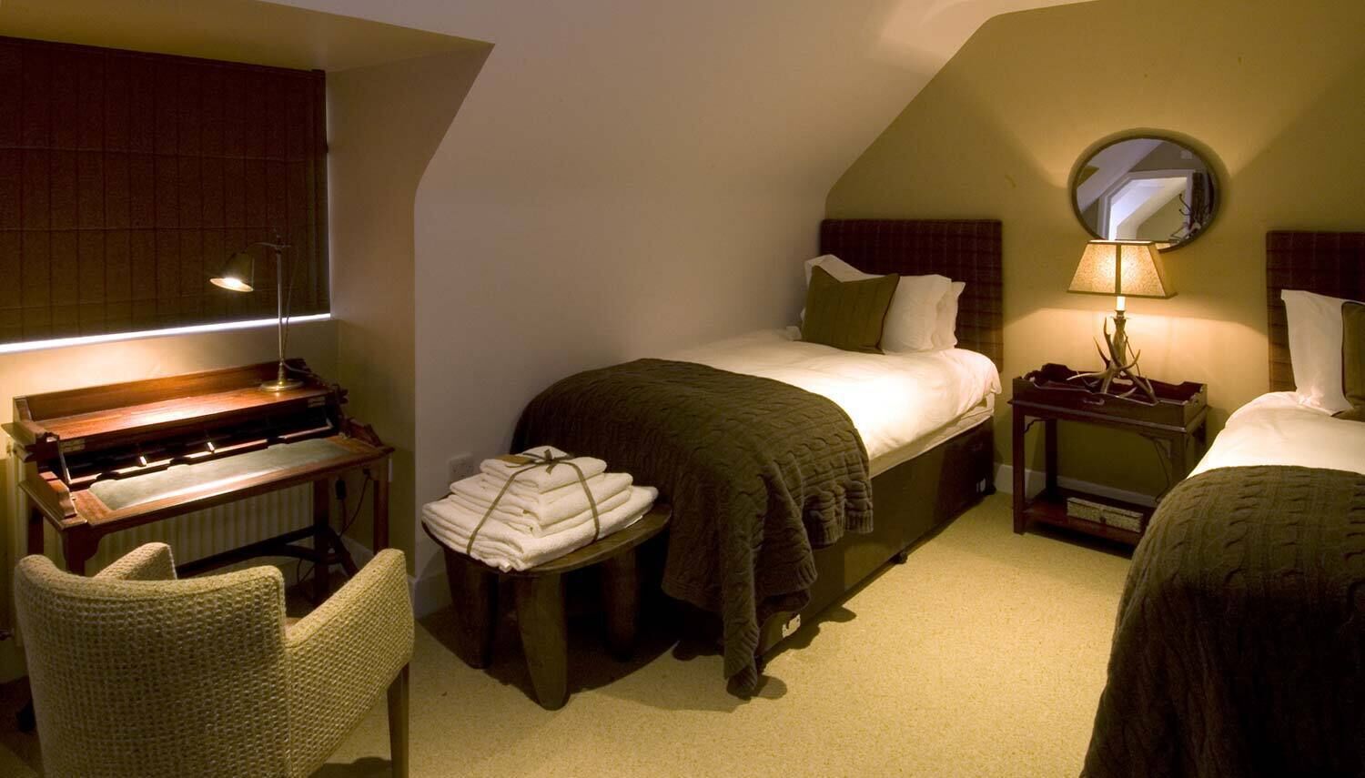 A cozy bedroom with a piano, a bed with a green blanket, a bedside table with a lamp, and a chair.