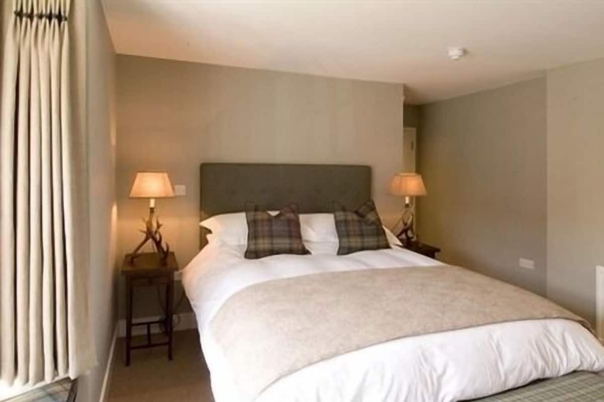 A neatly made bed with a headboard, two bedside lamps, and a window with curtains.