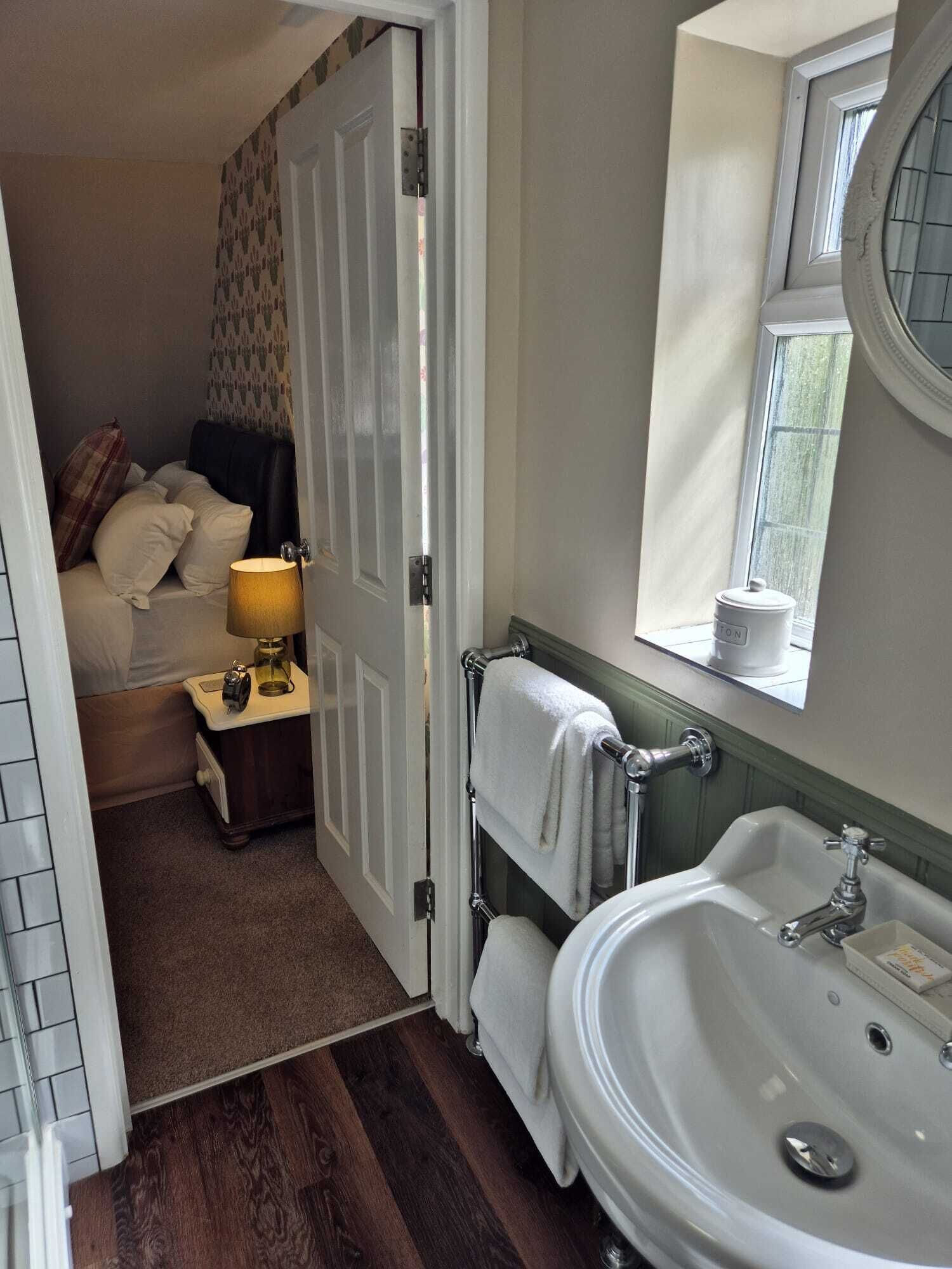 A bedroom with a bed, a bedside lamp, and a nightstand. There is a bathroom with a sink, a mirror, and a window.