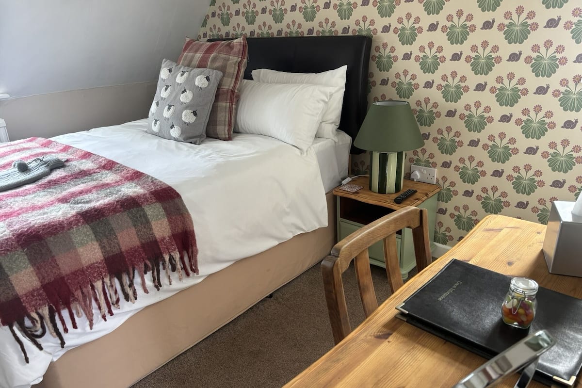 A neatly made bed with a plaid blanket, a bedside lamp, a wooden nightstand, and a small table with a remote control and a glass jar.