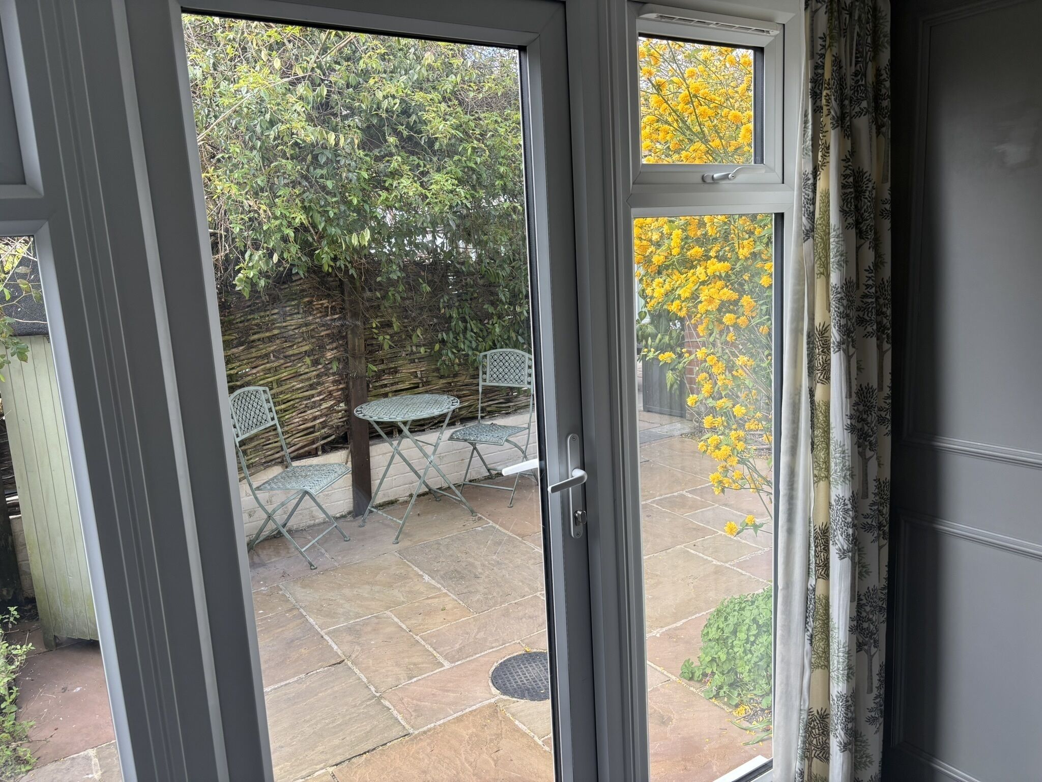 Un patio con una mesa y sillas, vistas a un jardín con flores amarillas, y una puerta entreabierta.