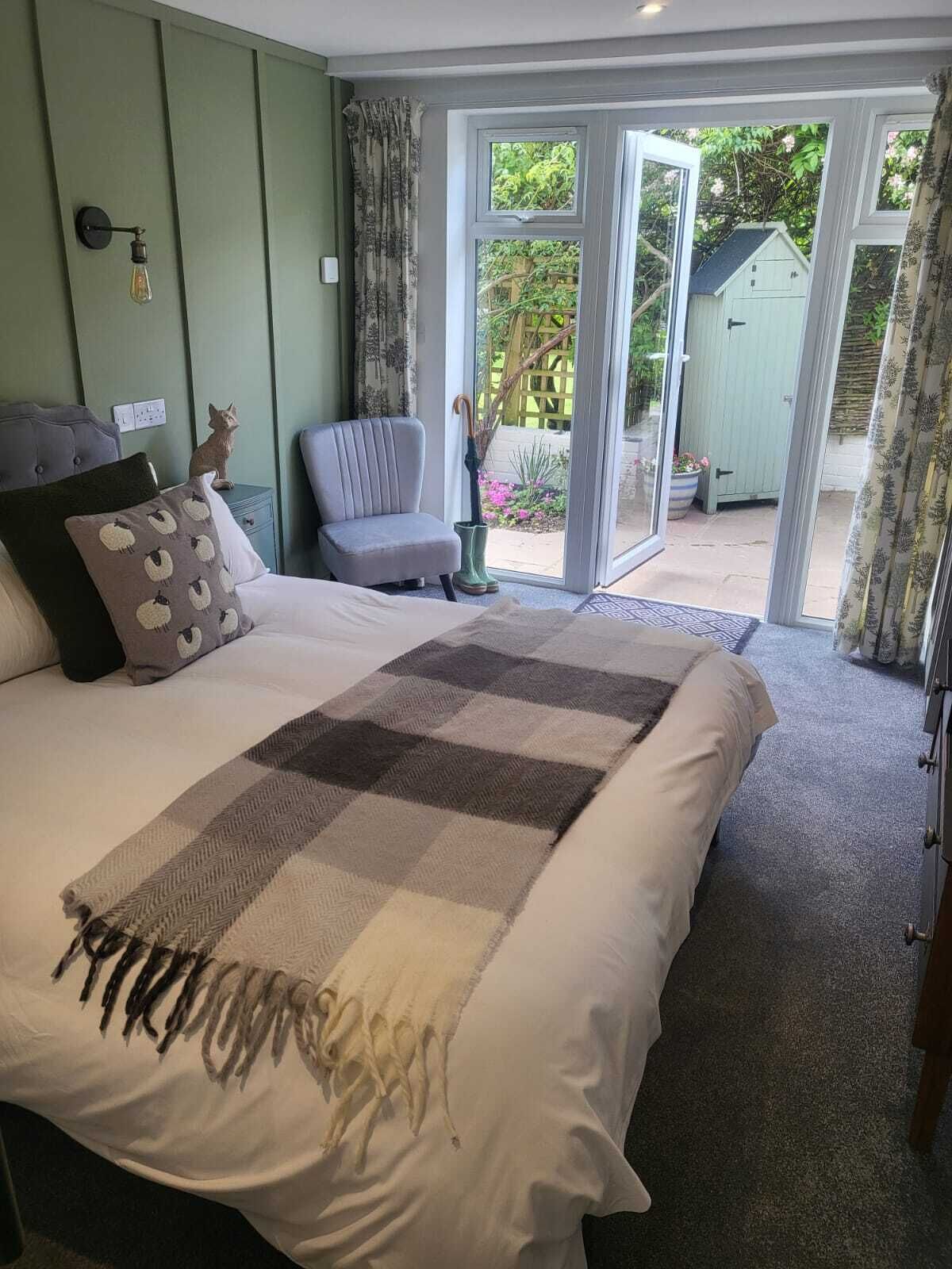 A bedroom with a large bed, a chair, a small table, and a window with a view of a garden.