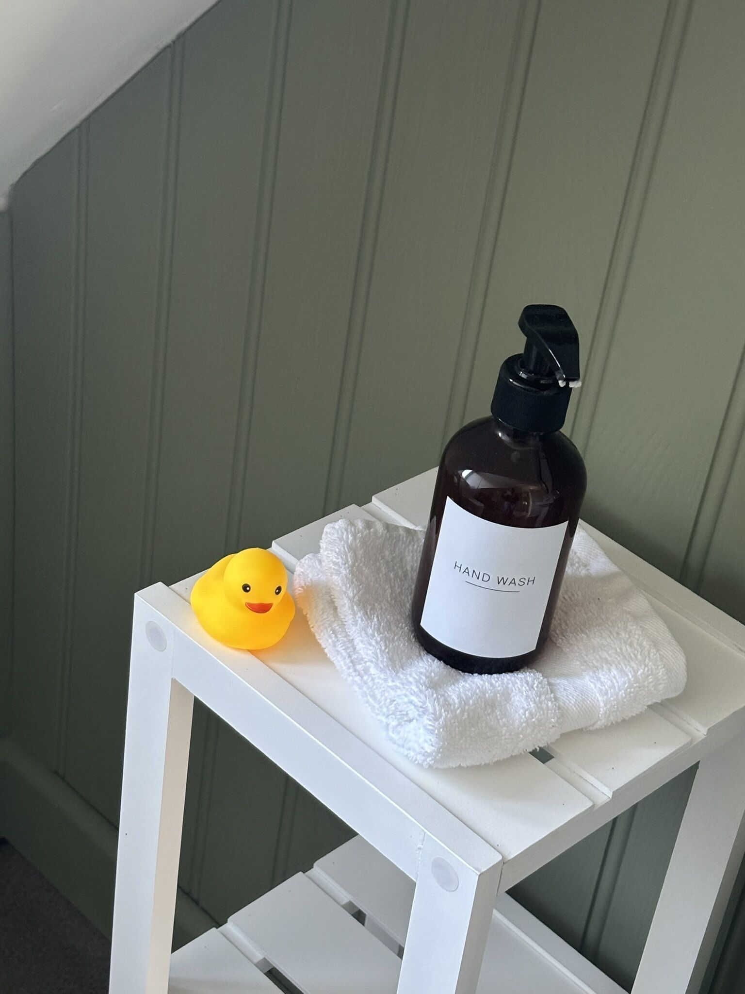 Una botella de jabón líquido para manos y un patito de goma sobre un estante blanco frente a una pared verde.