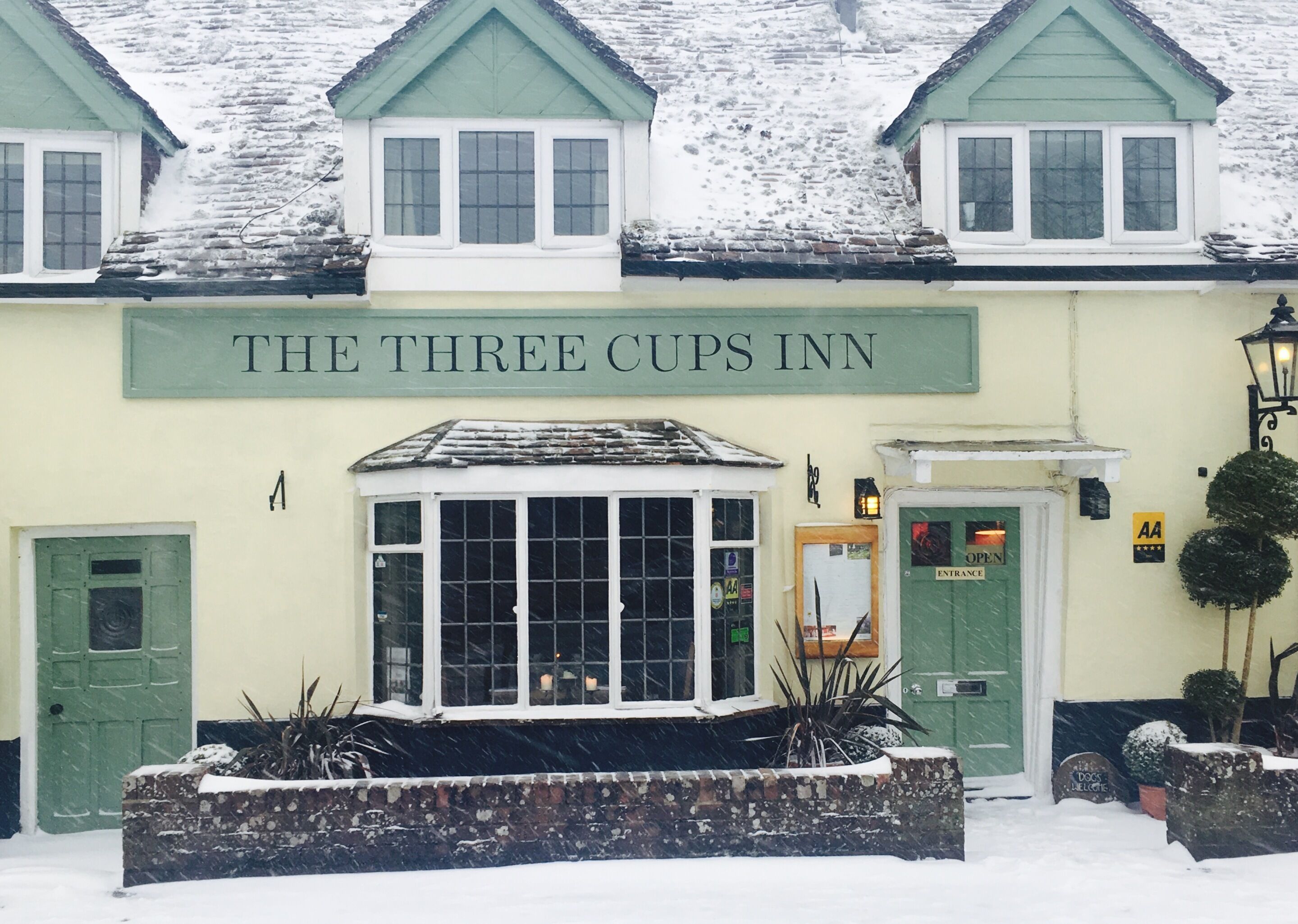 A building labeled "THE THREE CUPS INN" with a snow-covered roof, a green door, and a window with a view of the interior.