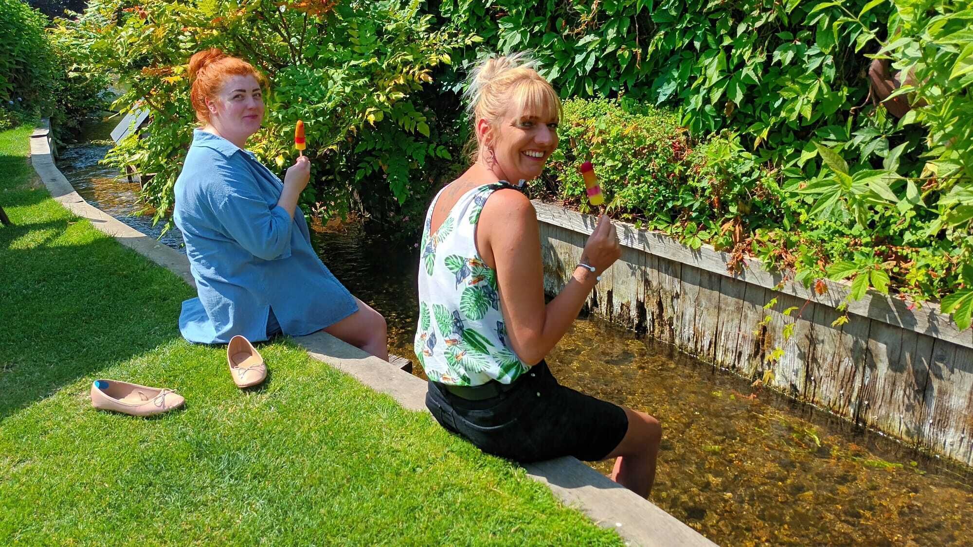 Deux personnes sont assises près d’un élément décoratif aquatique dans un jardin où la végétation est luxuriante.