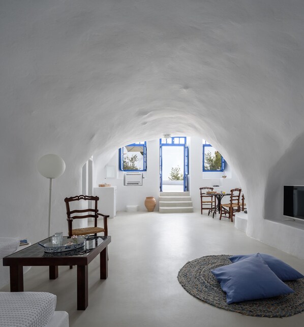 A spacious, white-walled interior with a high ceiling, a dining area with wooden furniture, and a view of the outdoors through blue-framed windows. - Aghios Artemios Traditional Houses (Santorini)