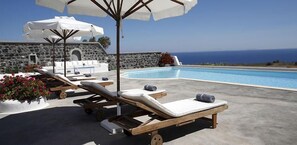 A poolside area with lounge chairs, umbrellas, and a stone wall backdrop. - Aghios Artemios Traditional Houses (Santorini)