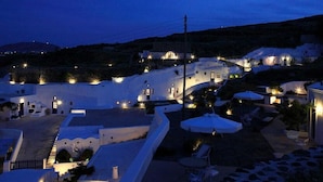 A coastal village at night with white-washed buildings, outdoor seating, and a hillside backdrop.
