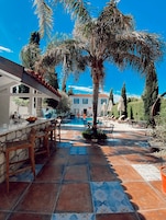 Zona junto a la piscina con un bar de piedra, sillas de madera y un patio con baldosas.