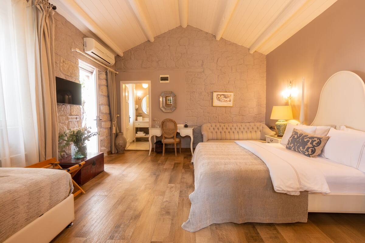 A bedroom with a large bed, a desk, and a chair. There is a dresser and a mirror. The room has wooden flooring and a stone wall.