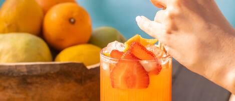 A tall glass with a fruit drink, a hand reaching for it, and a bowl of fruits in the background.