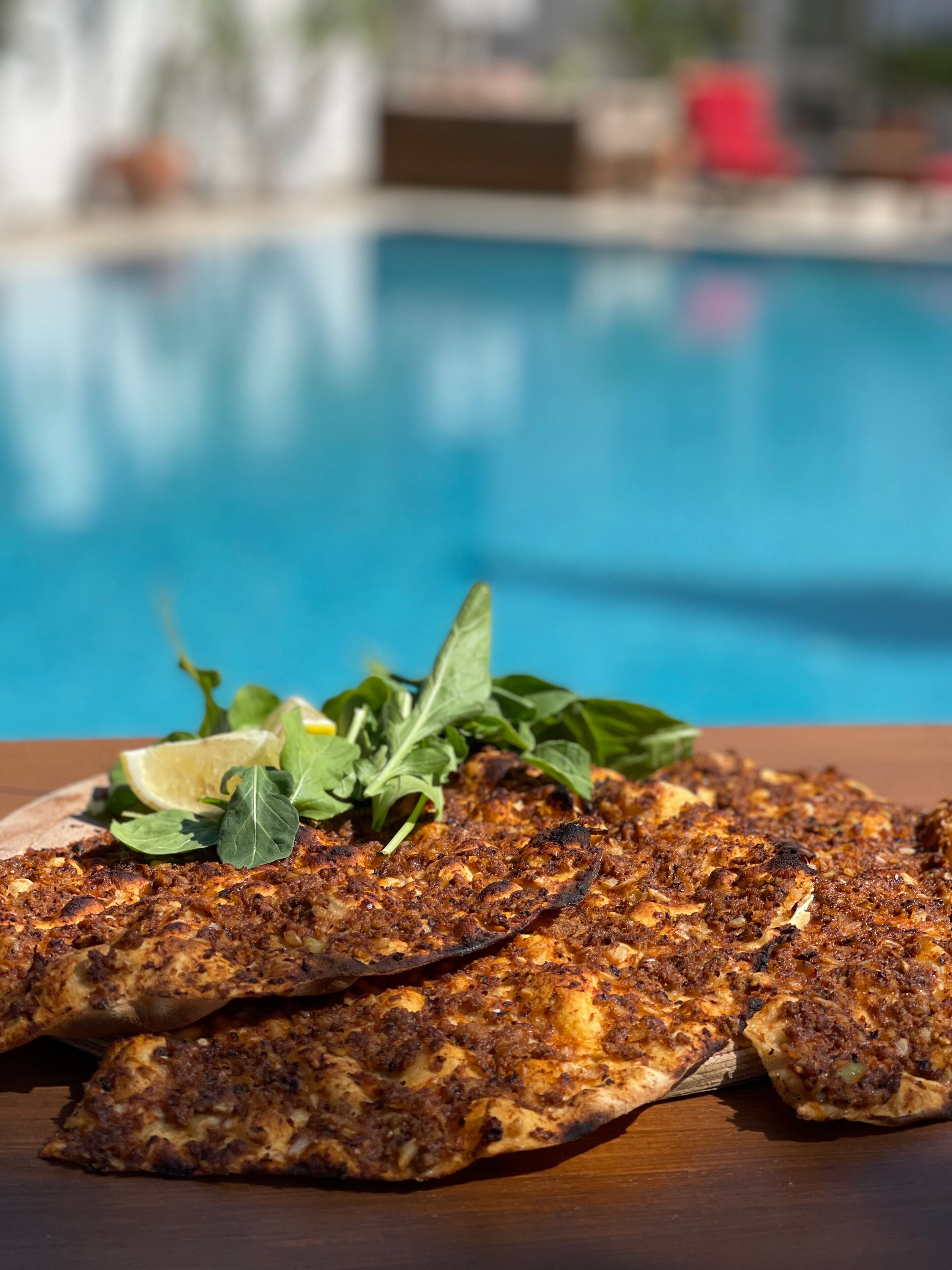 A grilled fish dish with lemon and greens on a wooden table by a pool.