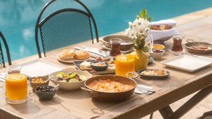 Une table dressée au bord de la piscine, avec des aliments pour le petit-déjeuner : un bol de céréales, un verre de jus d’orange et une assiette de viennoiseries.