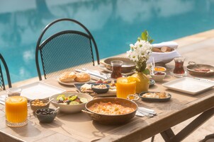 A table set with breakfast items by a poolside, including a bowl of cereal, a glass of orange juice, and a plate of pastries.