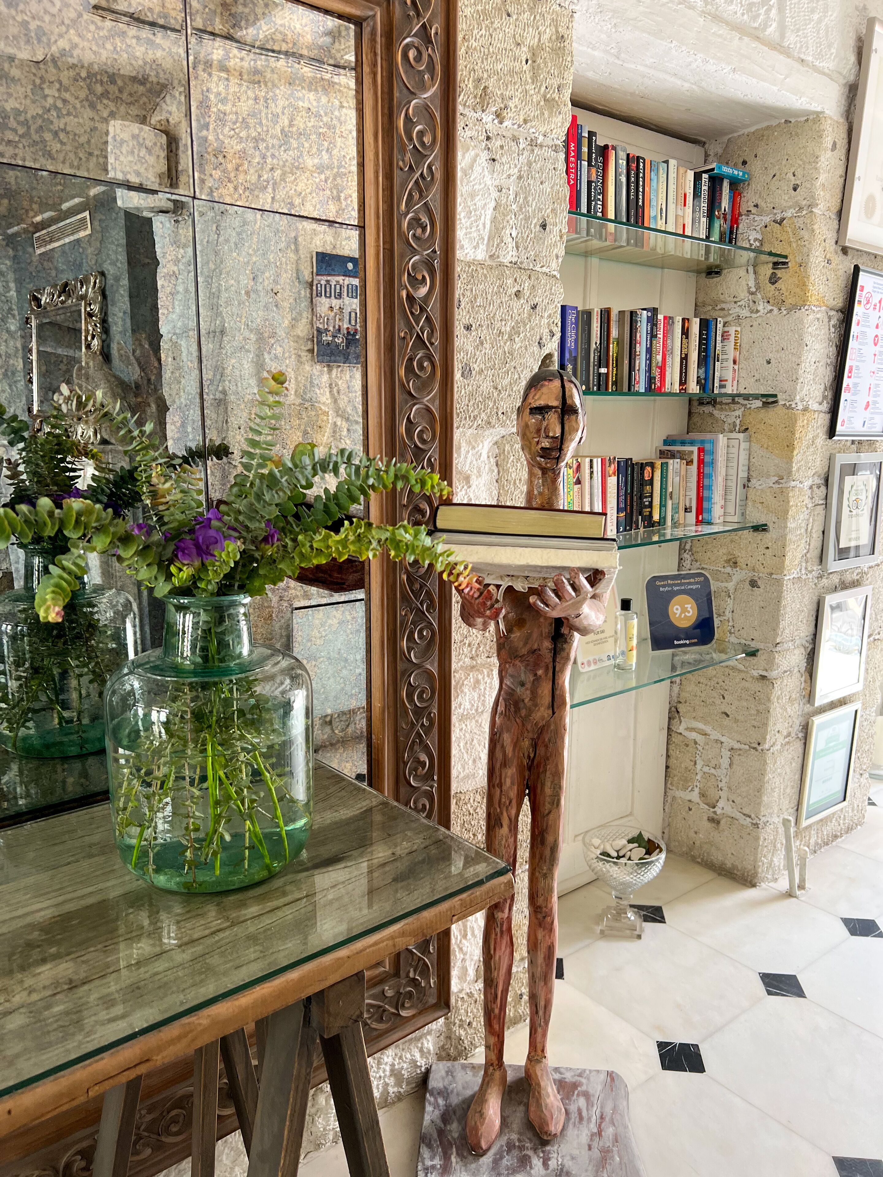 A statue holding a tray with a vase of flowers on a glass table, surrounded by bookshelves and a stone wall.