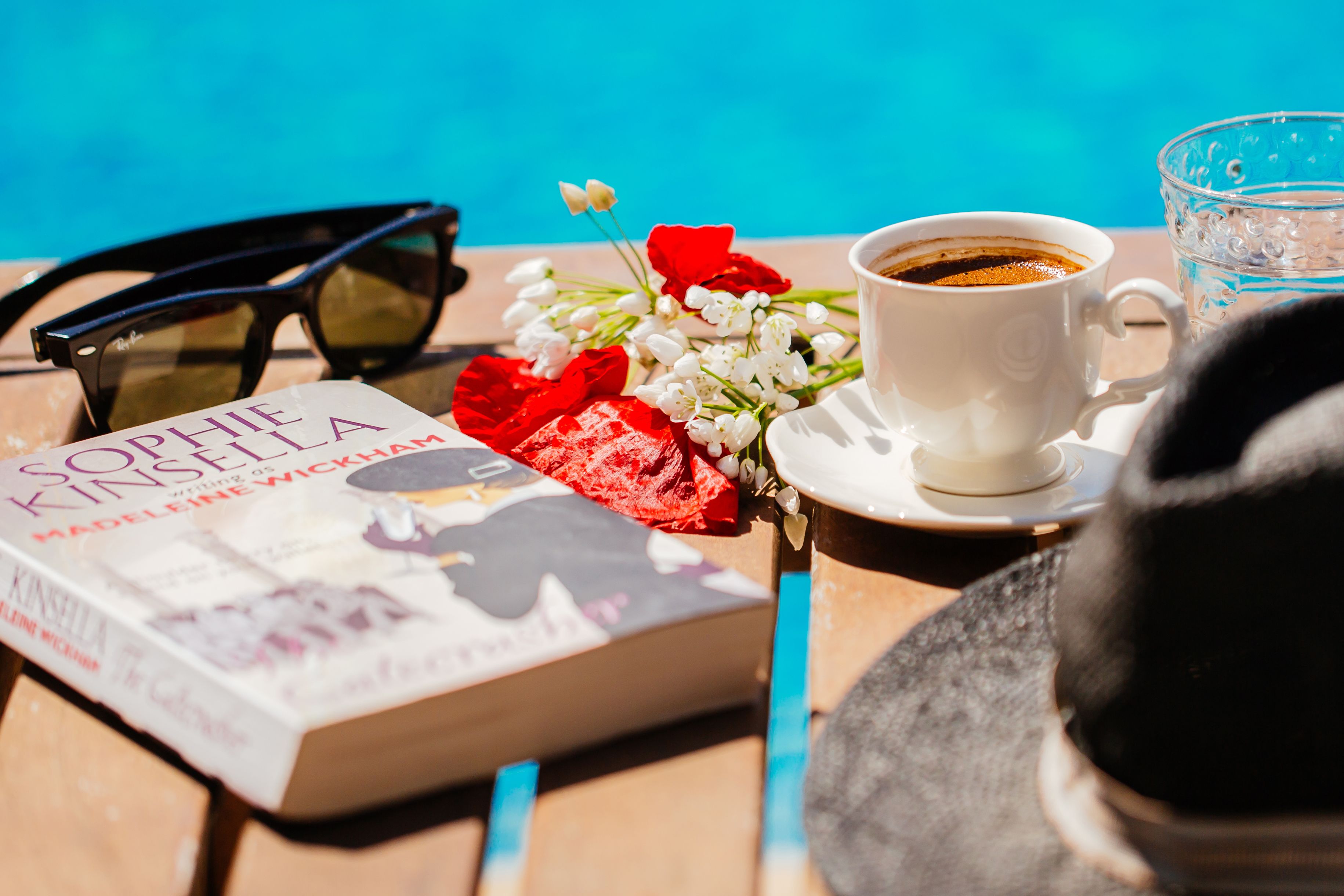 Un llibre anomenat 'Sophie Kinsella' de Madeleine Wickham, una tassa de cafè, ulleres de sol i un got d'aigua sobre una taula.
