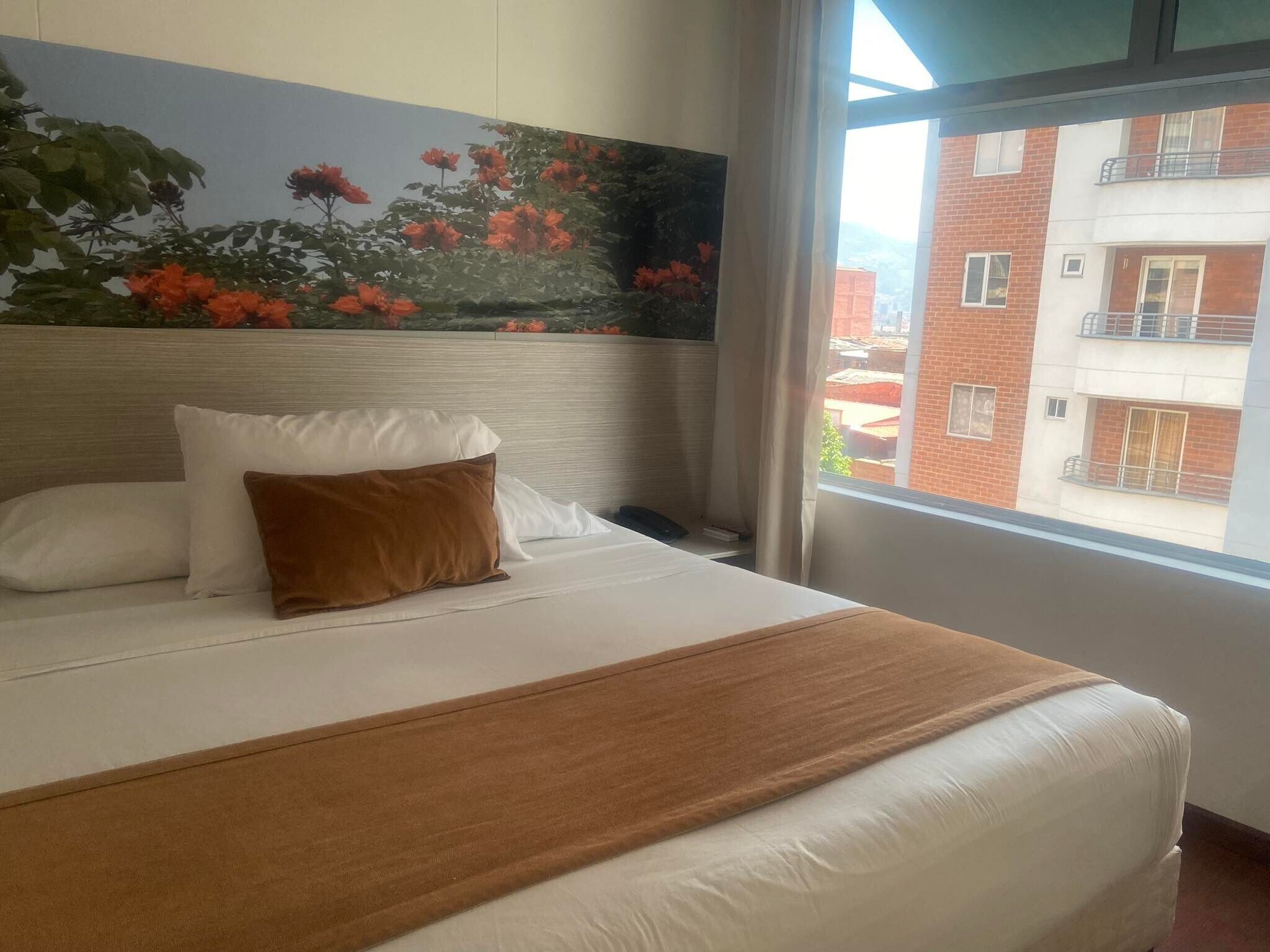 A hotel room with a bed, a painting of flowers on the wall, and a view of buildings outside the window.