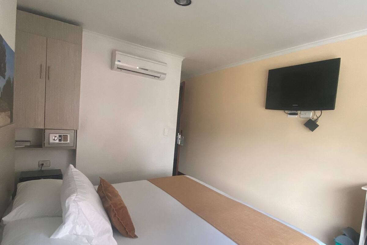 A hotel room with a bed, an air conditioning unit, a television mounted on the wall, and a safe.