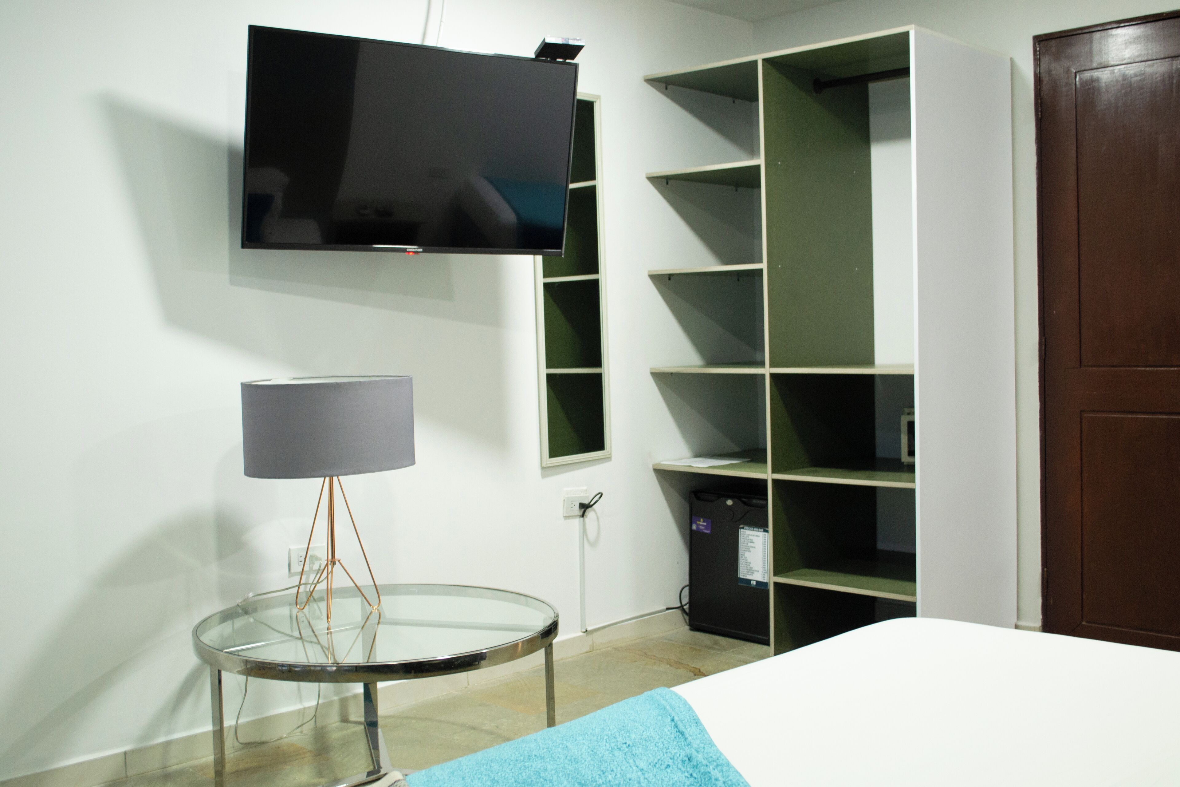 Ein Zimmer mit einem Bett, einem an der Wand montierten Fernseher, einer Lampe auf einem Nachttisch und einem Regal.