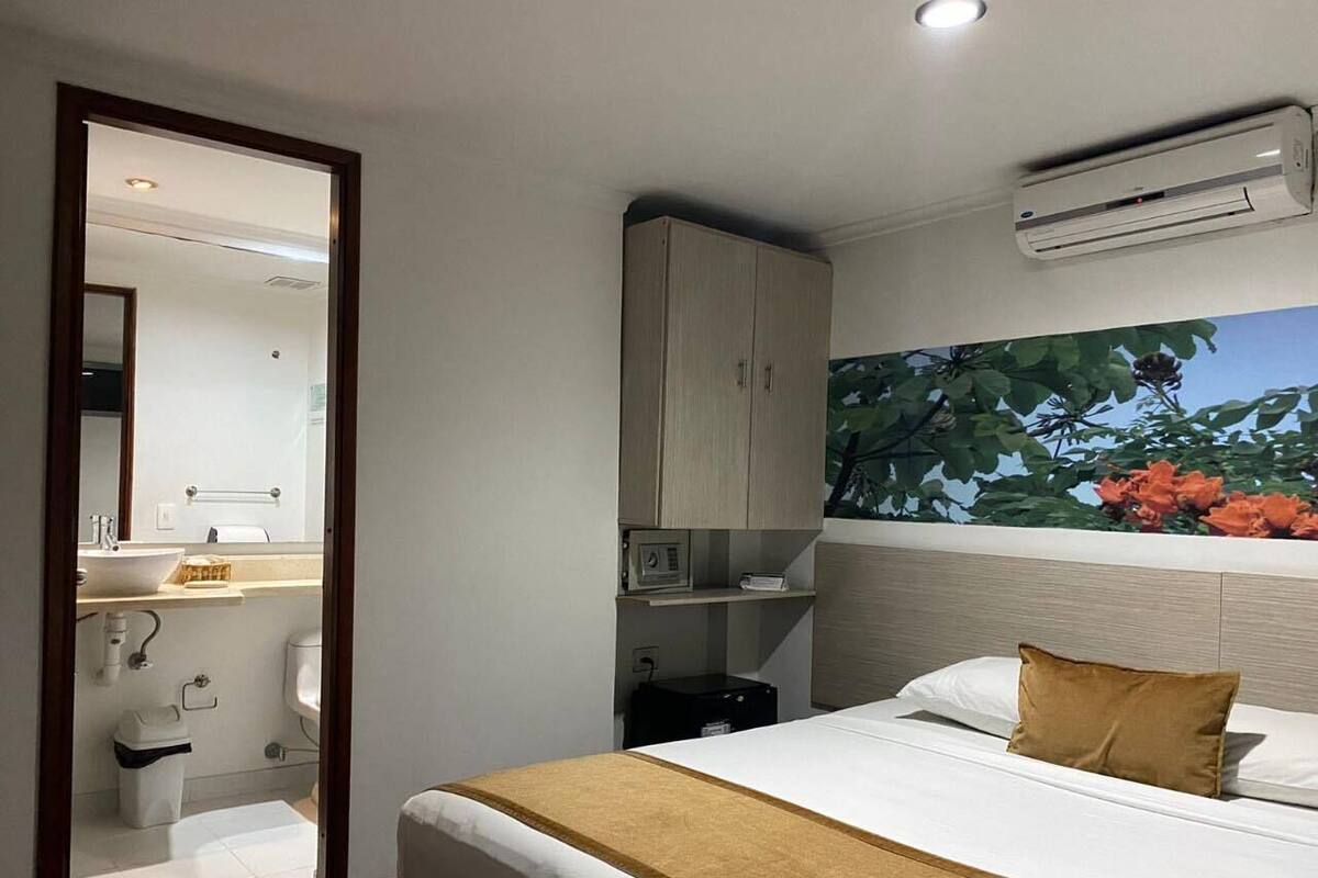 A hotel room with a bed, a nightstand, a wardrobe, a bathroom visible through an open door, and a wall-mounted air conditioner.