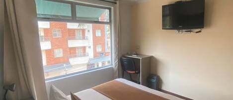 A hotel room with a bed, a desk, a chair, a television, and a window with a view of a building.