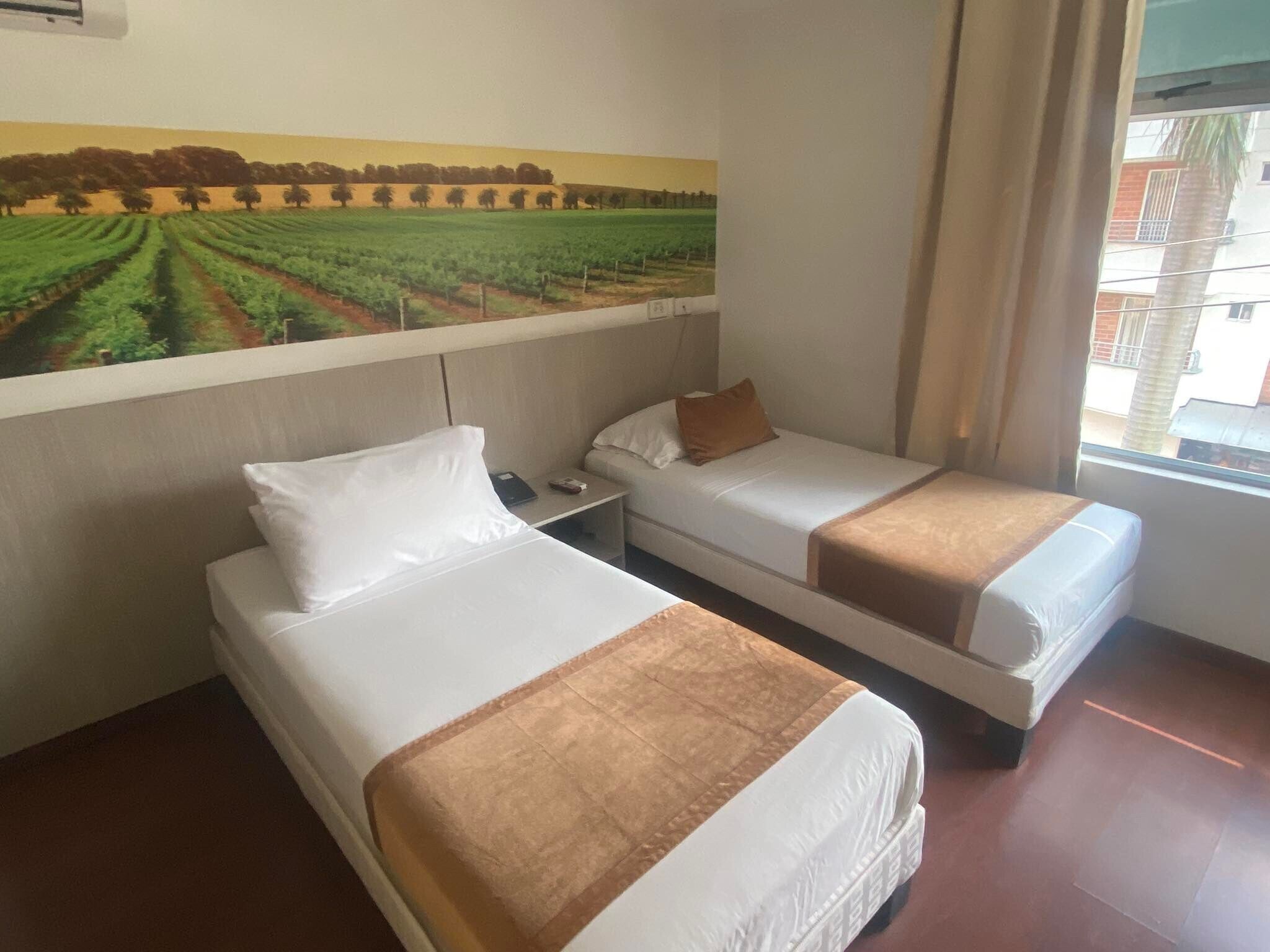 A hotel room with two beds, a nightstand, a painting of a field, and a window with curtains.