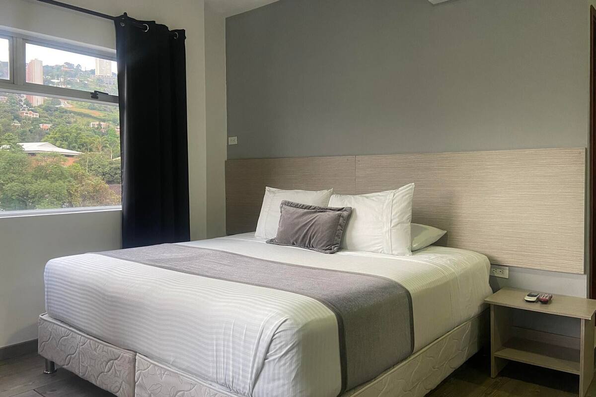 A neatly made bed with a headboard, a bedside table with a phone, and a window with a view of buildings and trees.