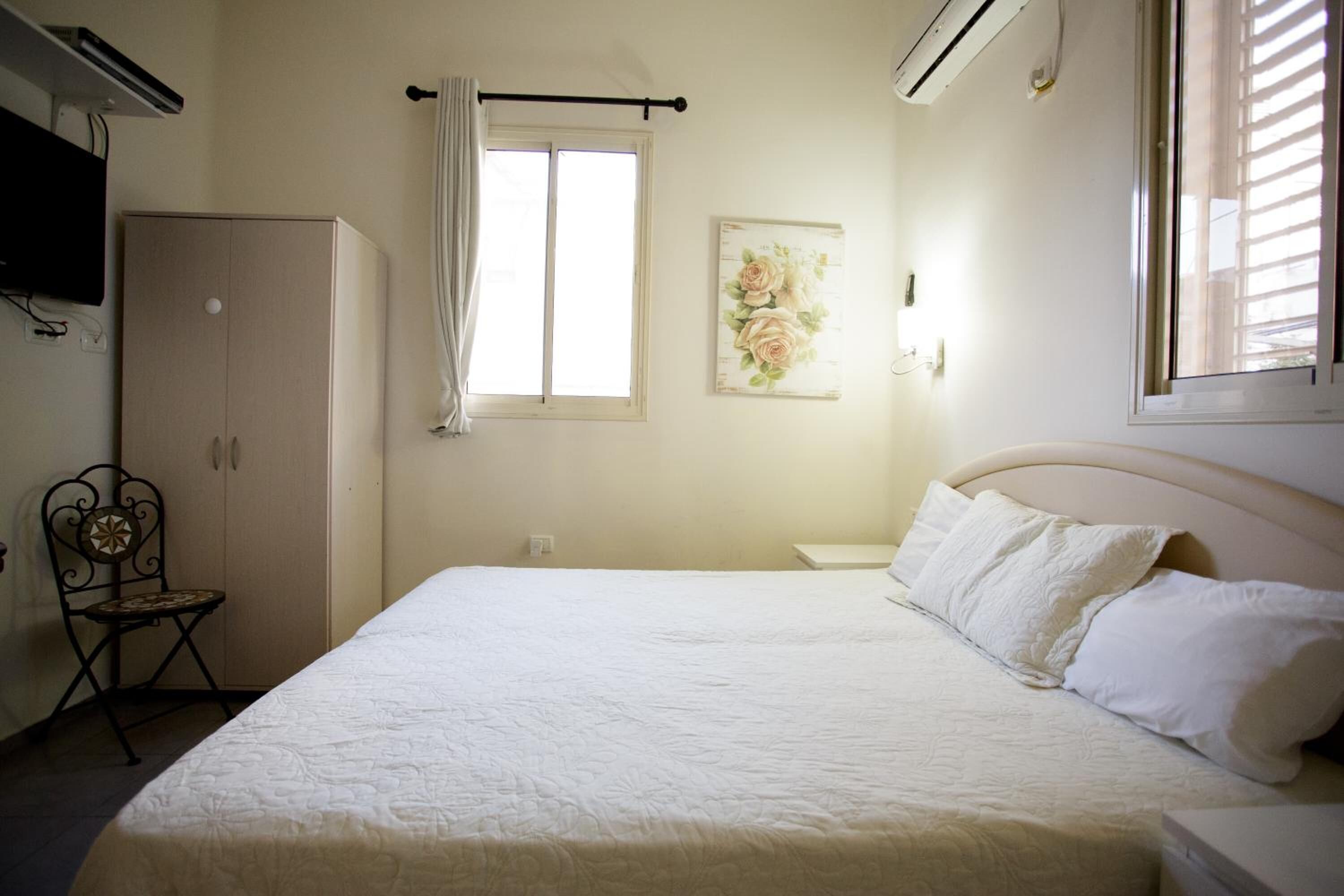 Un dormitorio con cama, armario, silla, ventana con cortinas y un cuadro colgado en la pared.