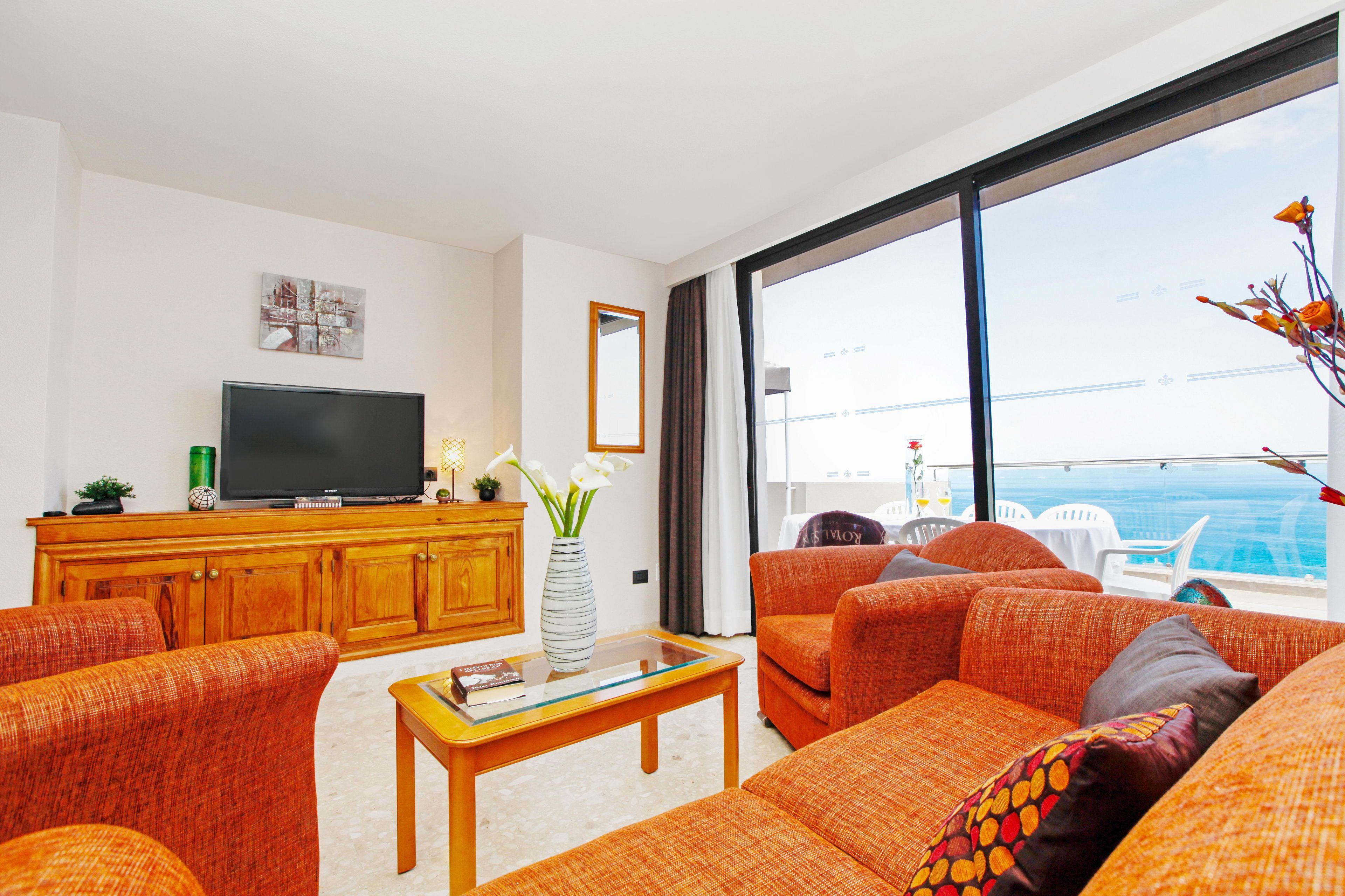 A modern living room with a flat-screen TV, orange sofas, a wooden cabinet, and a large window offering a view of the ocean.