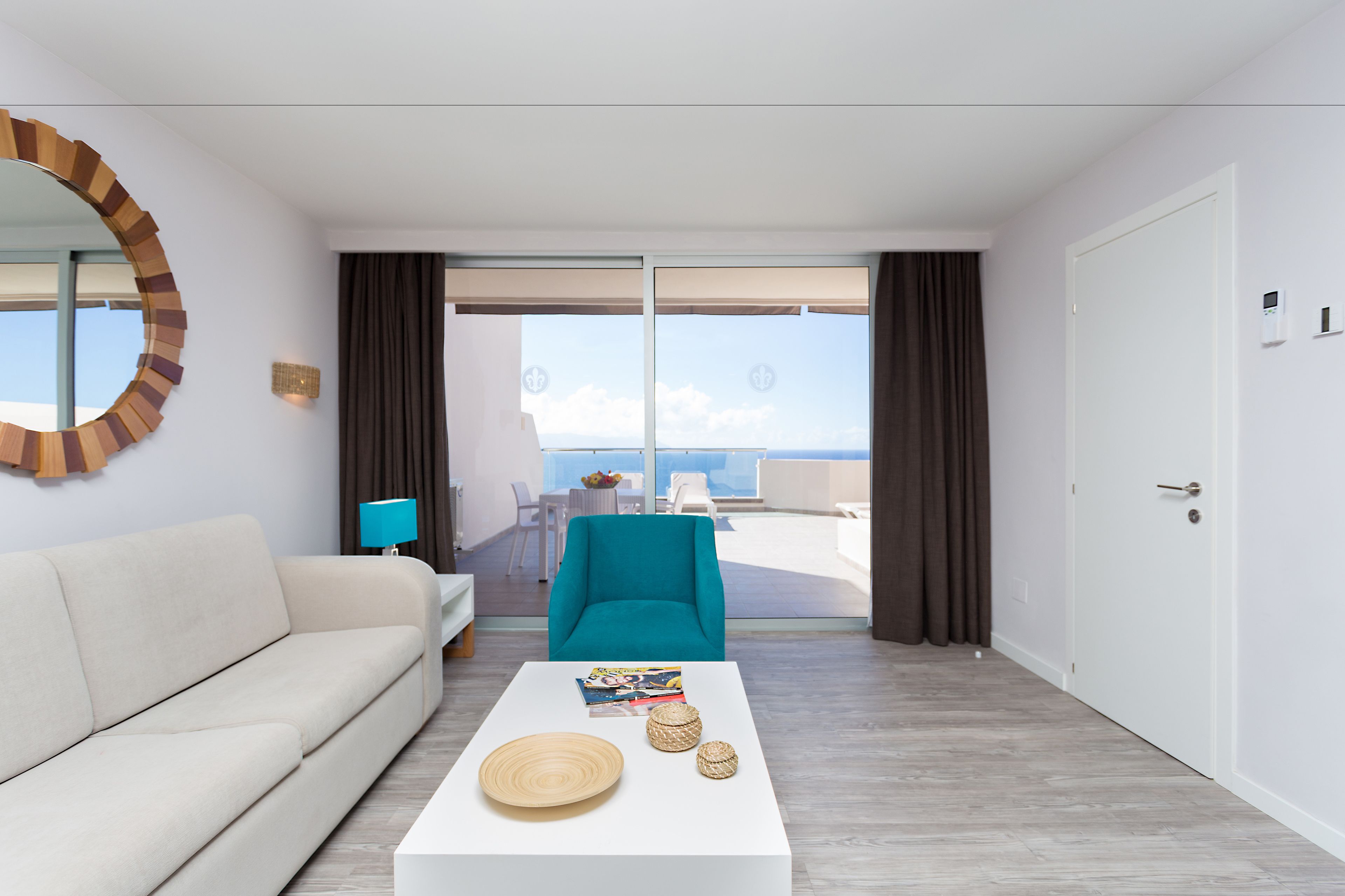 Una habitación de hotel moderna con un sofá blanco, un sillón azul verdoso, una mesita de café blanca con adornos y un ventanal con vistas al mar.
