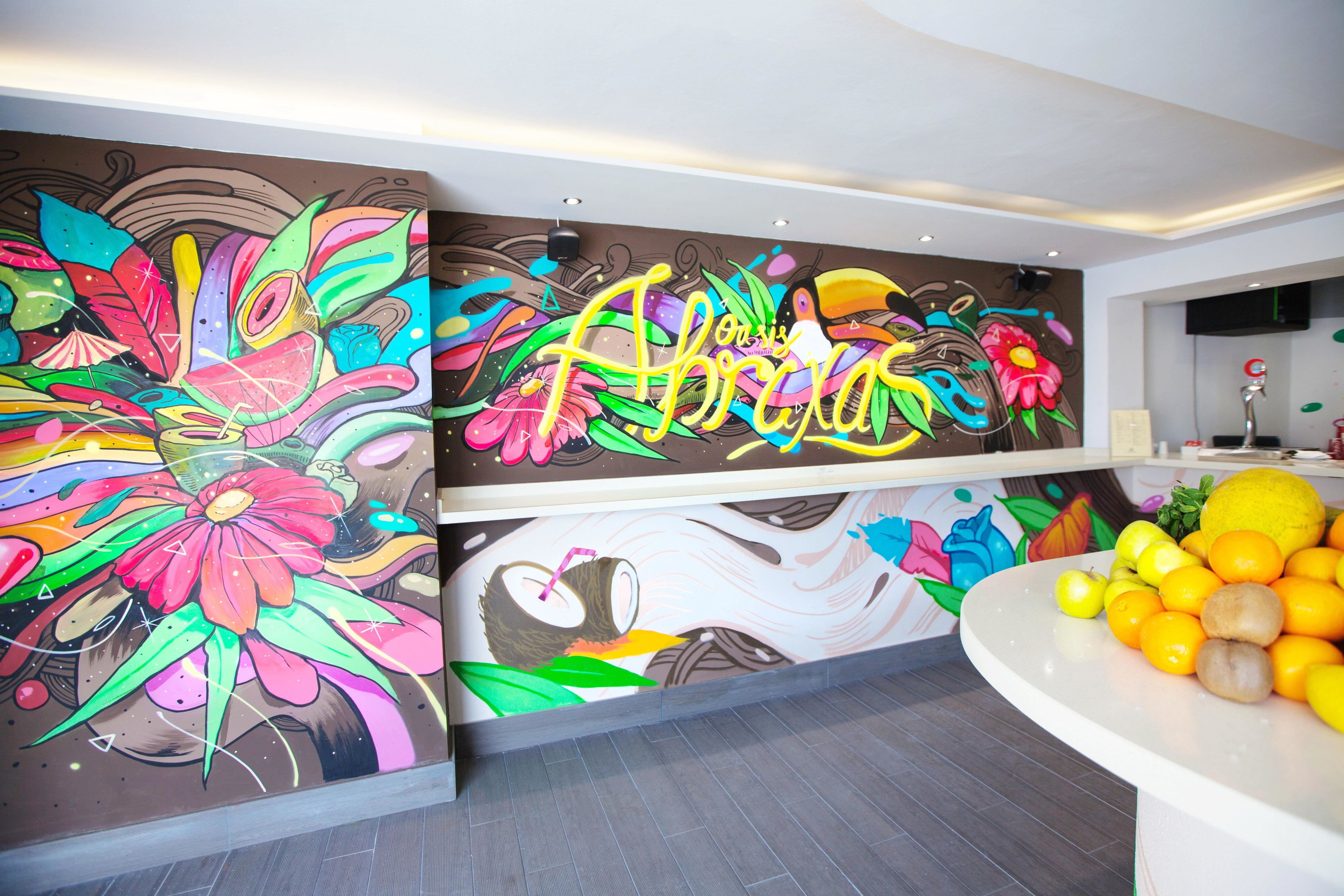 Un mural vibrante con diseños florales y abstractos, un mostrador de recepción blanco con detalles en naranja y un televisor de pantalla plana montado en la pared.