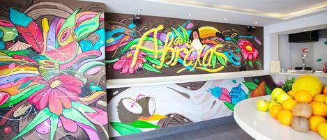 Un mural vibrante con diseños florales y abstractos, un mostrador de recepción blanco con detalles en naranja y un televisor de pantalla plana montado en la pared.
