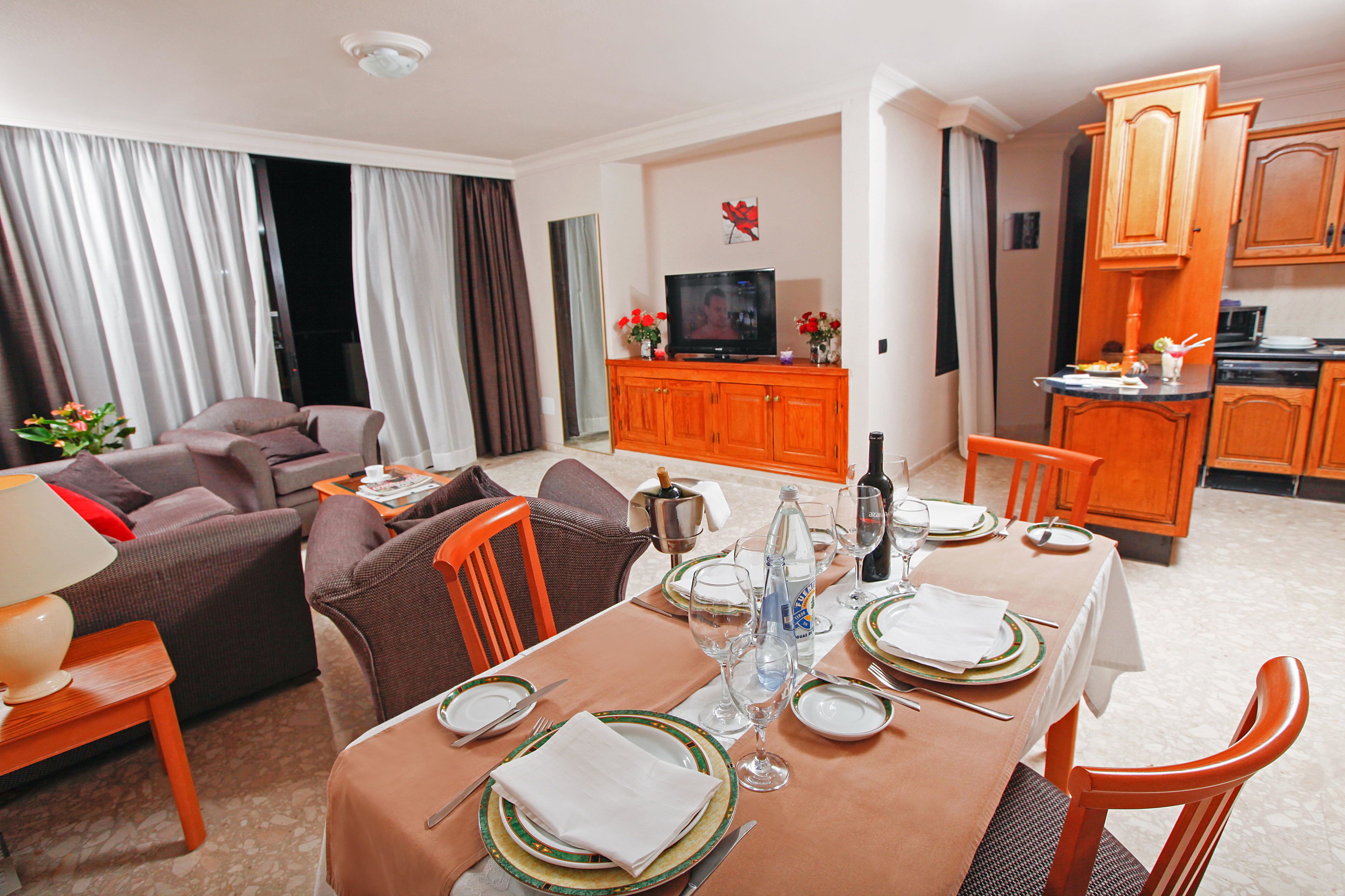 A well-lit living room with a sofa, dining table set for four, and a kitchen area with wooden cabinets.