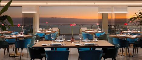 Un restaurant où les tables sont dressées pour le service, de grandes fenêtres offrant une vue sur l’océan et un décor intérieur moderne.
