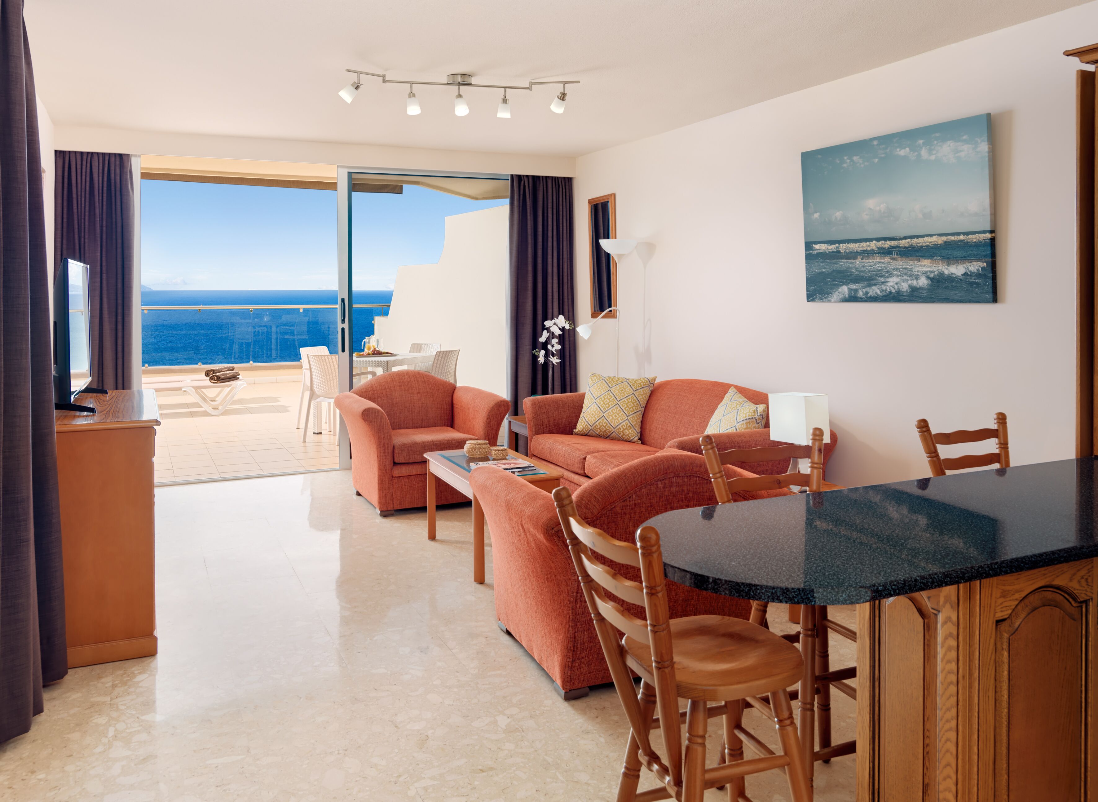 A modern living room with a view of the ocean, a dining area, and a balcony with a table and chairs.