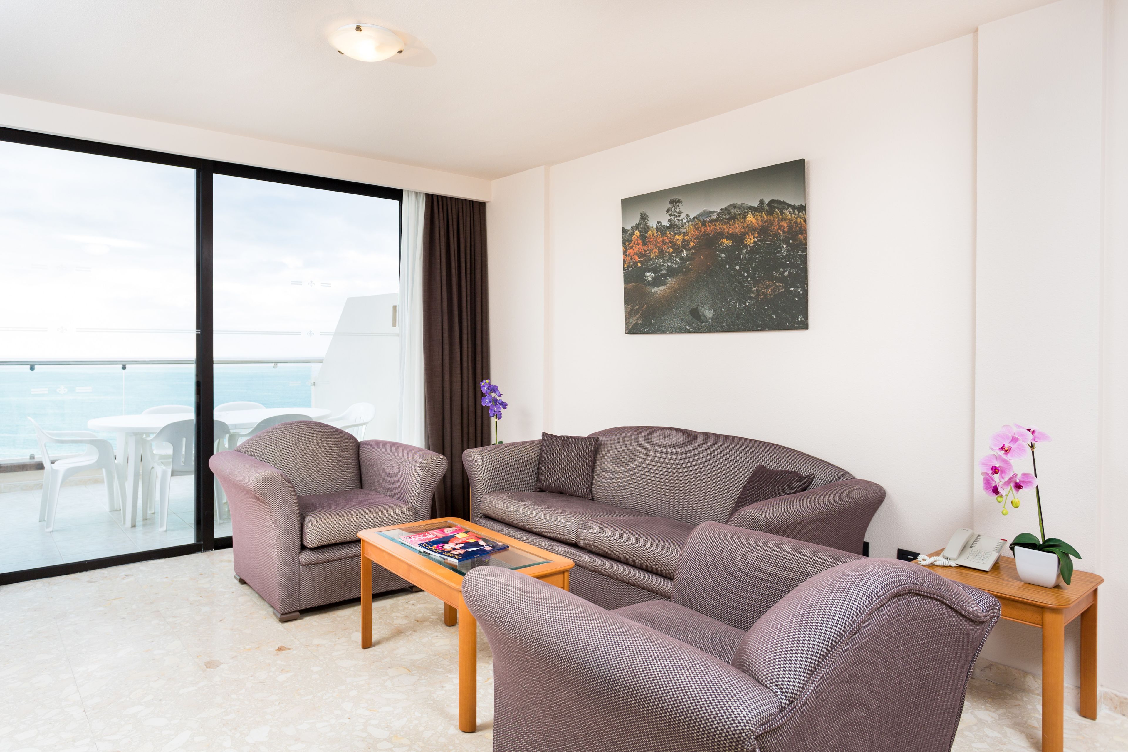 A modern living room with a sofa, armchairs, a coffee table, and a view of the sea through large windows.