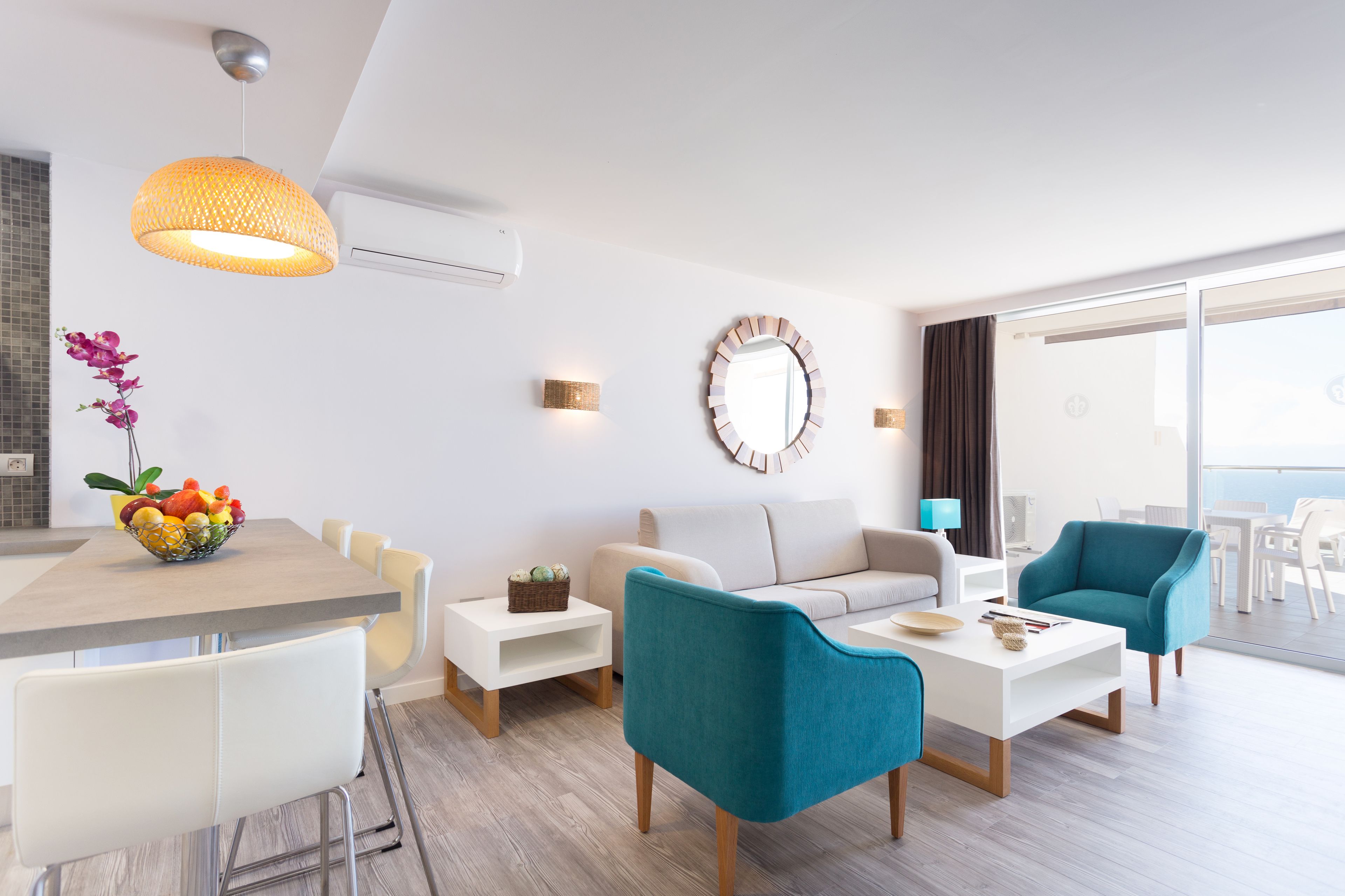 A modern living room with a dining area, a sofa, two armchairs, a coffee table, a fruit bowl, a hanging light fixture, a wall-mounted mirror, and a sliding glass door leading to a balcony.