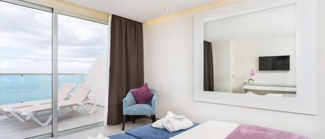 A hotel room with a bed, a large window with a view of the sea, a chair, and a TV.