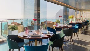 Un restaurante con mesas redondas, sillas azules y ventanas amplias que ofrecen vistas al mar.