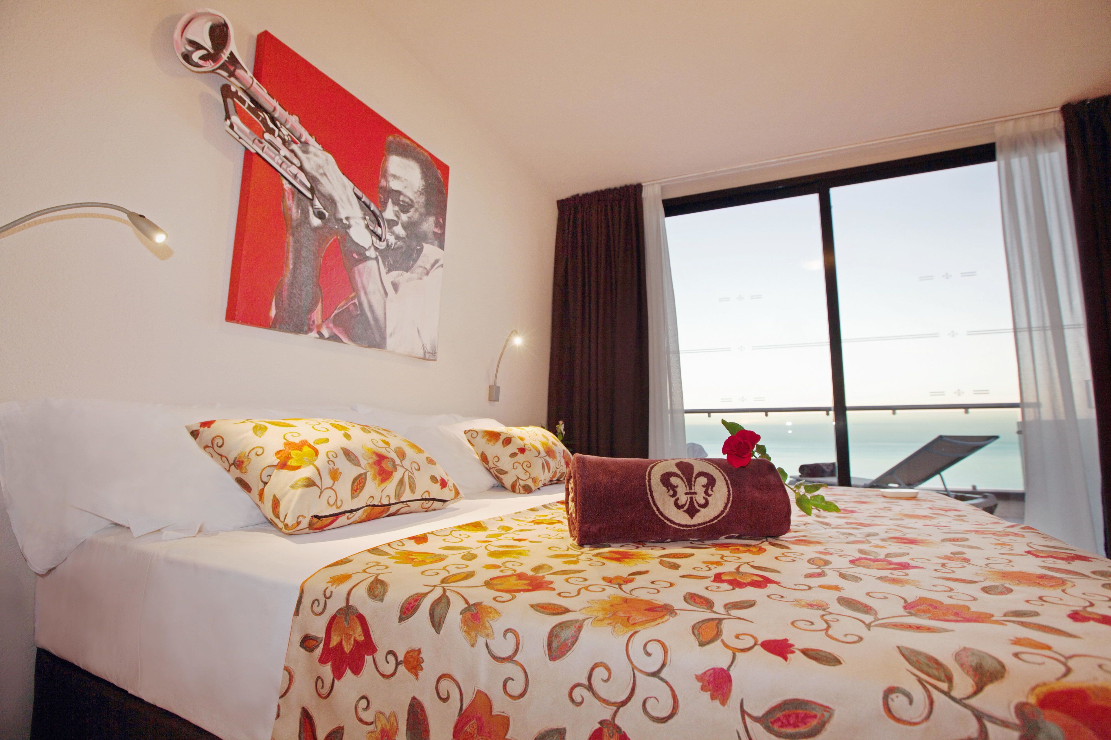 Habitación de hotel con cama, colcha estampada, un cuadro rojo y blanco, ventana con vista al mar y una tumbona afuera.