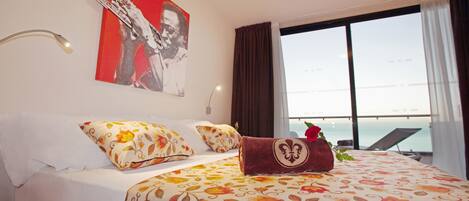 Habitación de hotel con cama, colcha estampada, un cuadro rojo y blanco, ventana con vista al mar y una tumbona afuera.