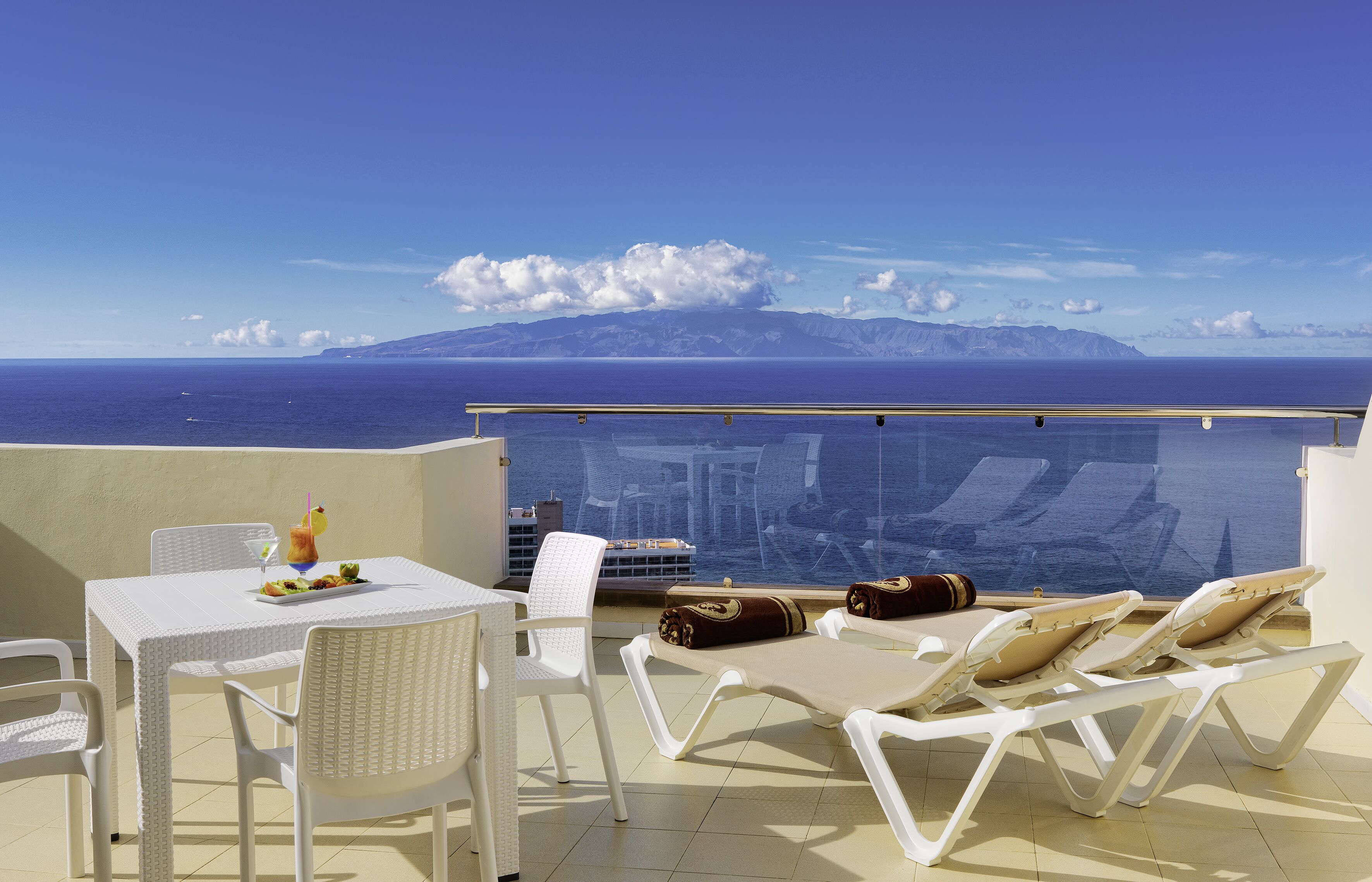 Ein Balkon mit weißen Stühlen und einem Tisch, Blick auf das Meer und die Berge, und ein klarer Himmel.