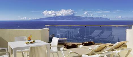 Ein Balkon mit weißen Stühlen und einem Tisch, Blick auf das Meer und die Berge, und ein klarer Himmel.