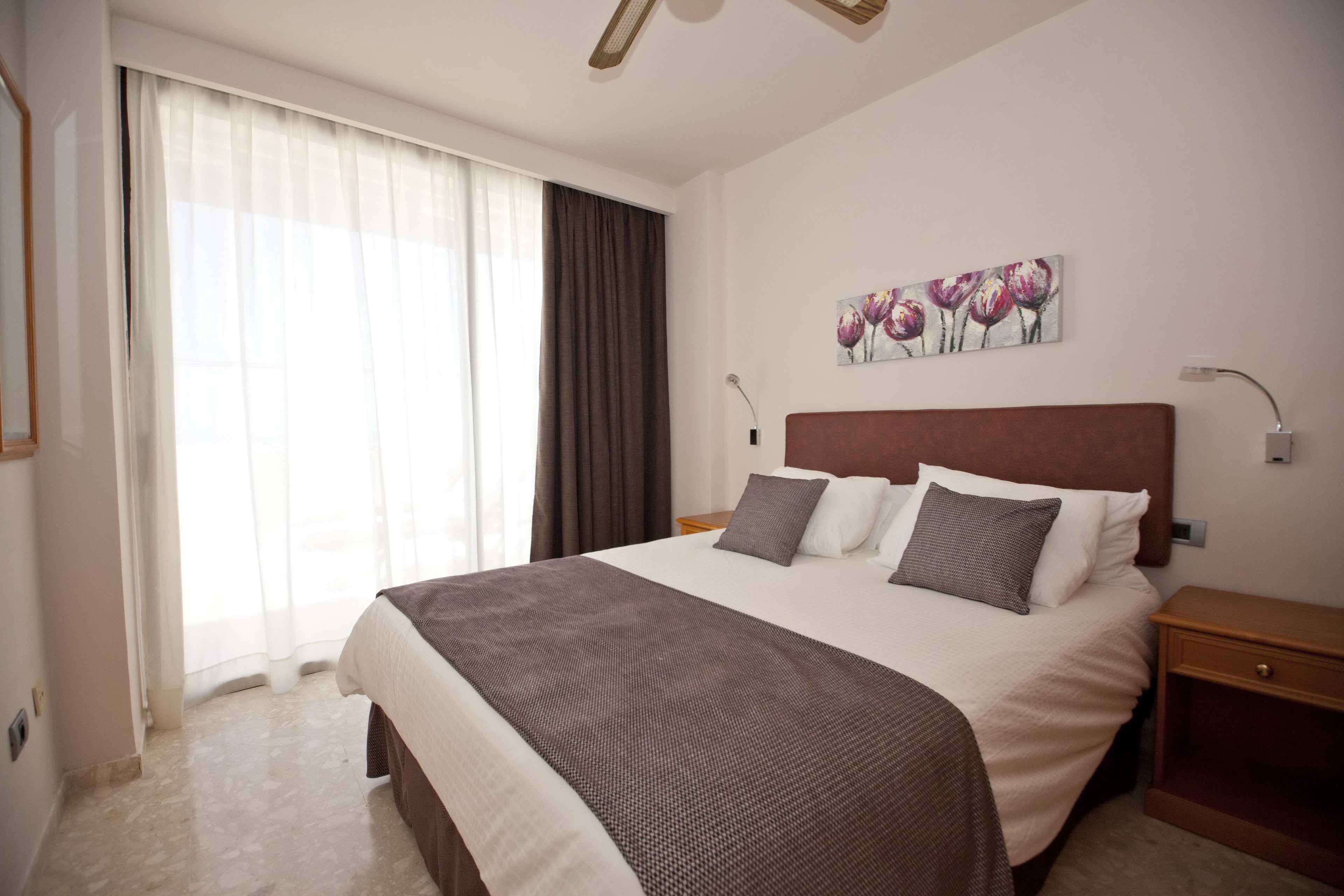 A hotel room with a large bed, a nightstand, a painting on the wall, and a window with curtains.