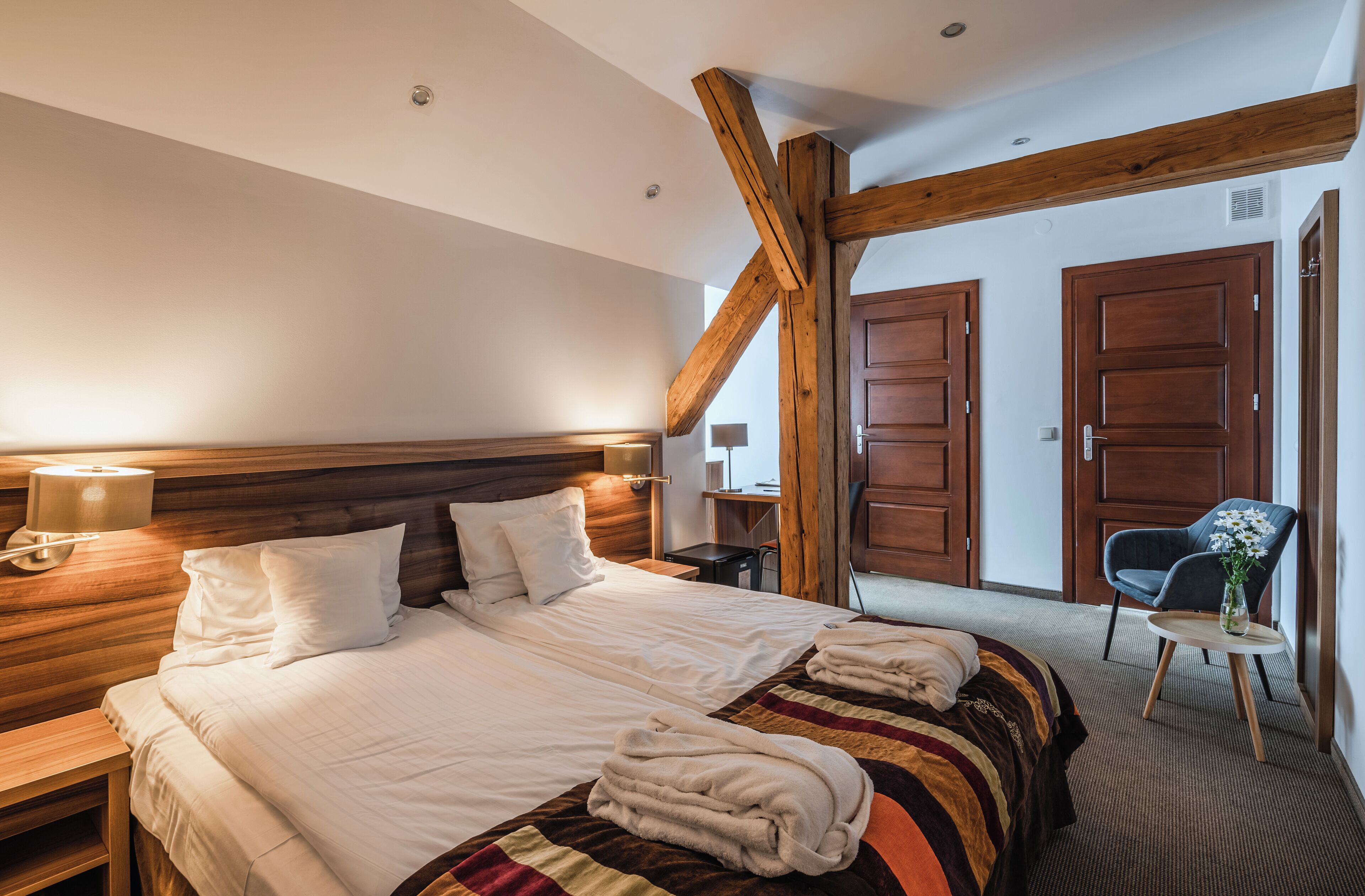 A hotel room with a large bed, wooden headboard, two bedside tables, a chair, and a small table with a vase.