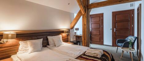 Una habitación de hotel con una cama grande, cabecera de madera, dos mesitas de noche, una silla y una pequeña mesa con un florero.