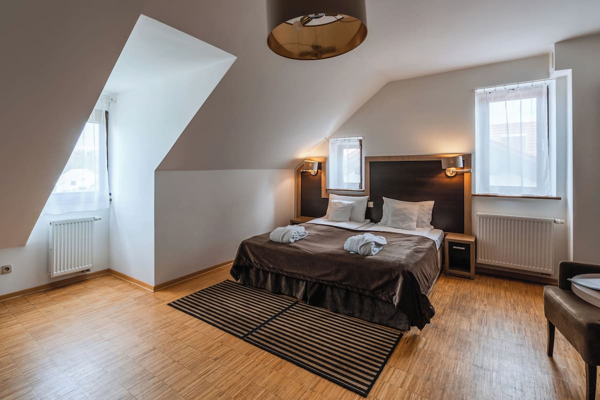 A bedroom with a sloped ceiling, a bed with a headboard, bedside lamps, a window, and a radiator.