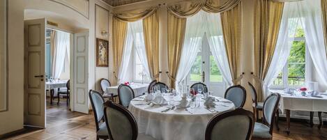 Comedor con una mesa redonda dispuesta para seis personas, rodeada por seis sillas. El ambiente tiene piso de madera, paredes color crema y ventanas amplias con cortinas translúcidas.
