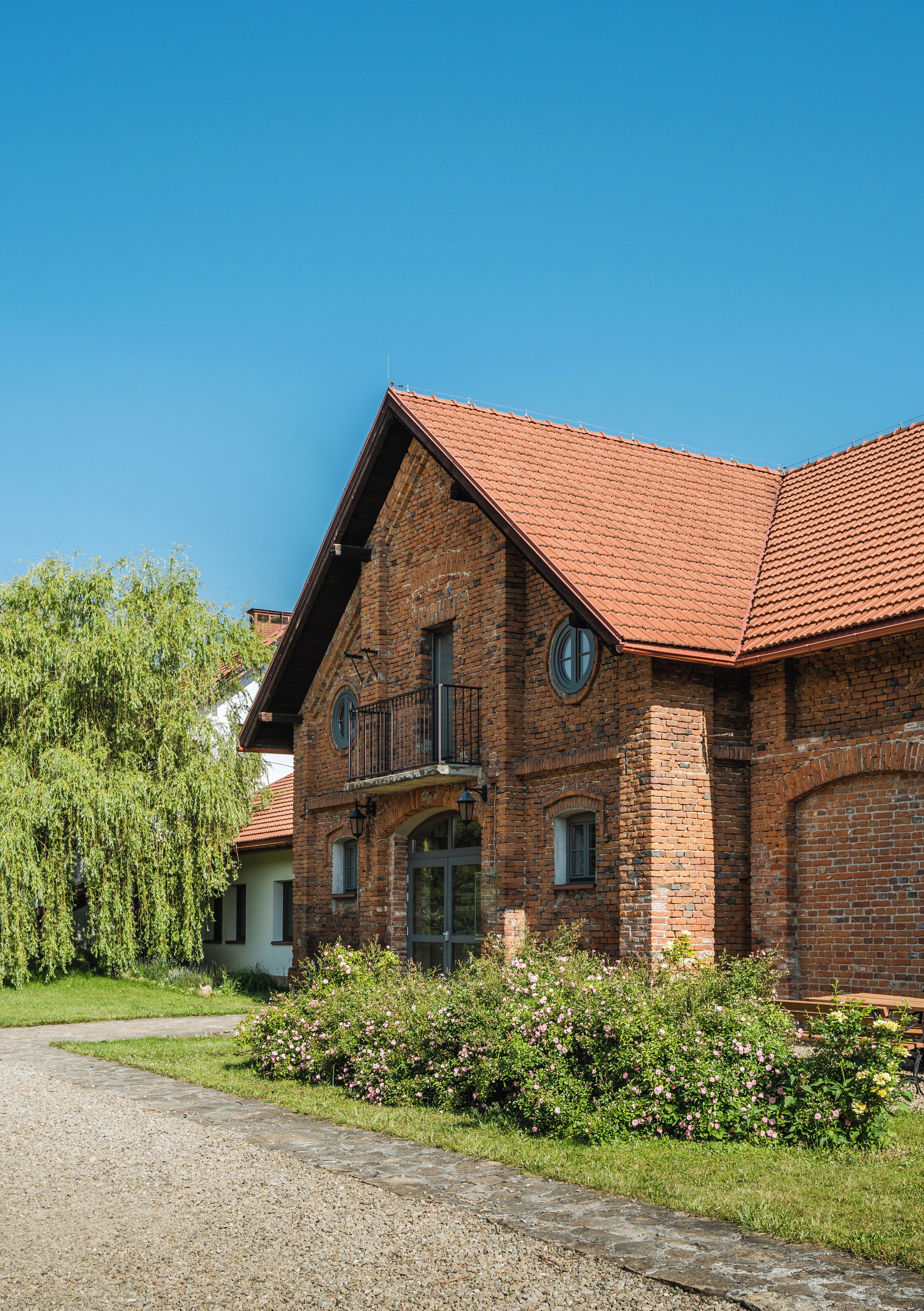 A brick building with a red-tiled roof, a balcony, and a garden with pink flowers.