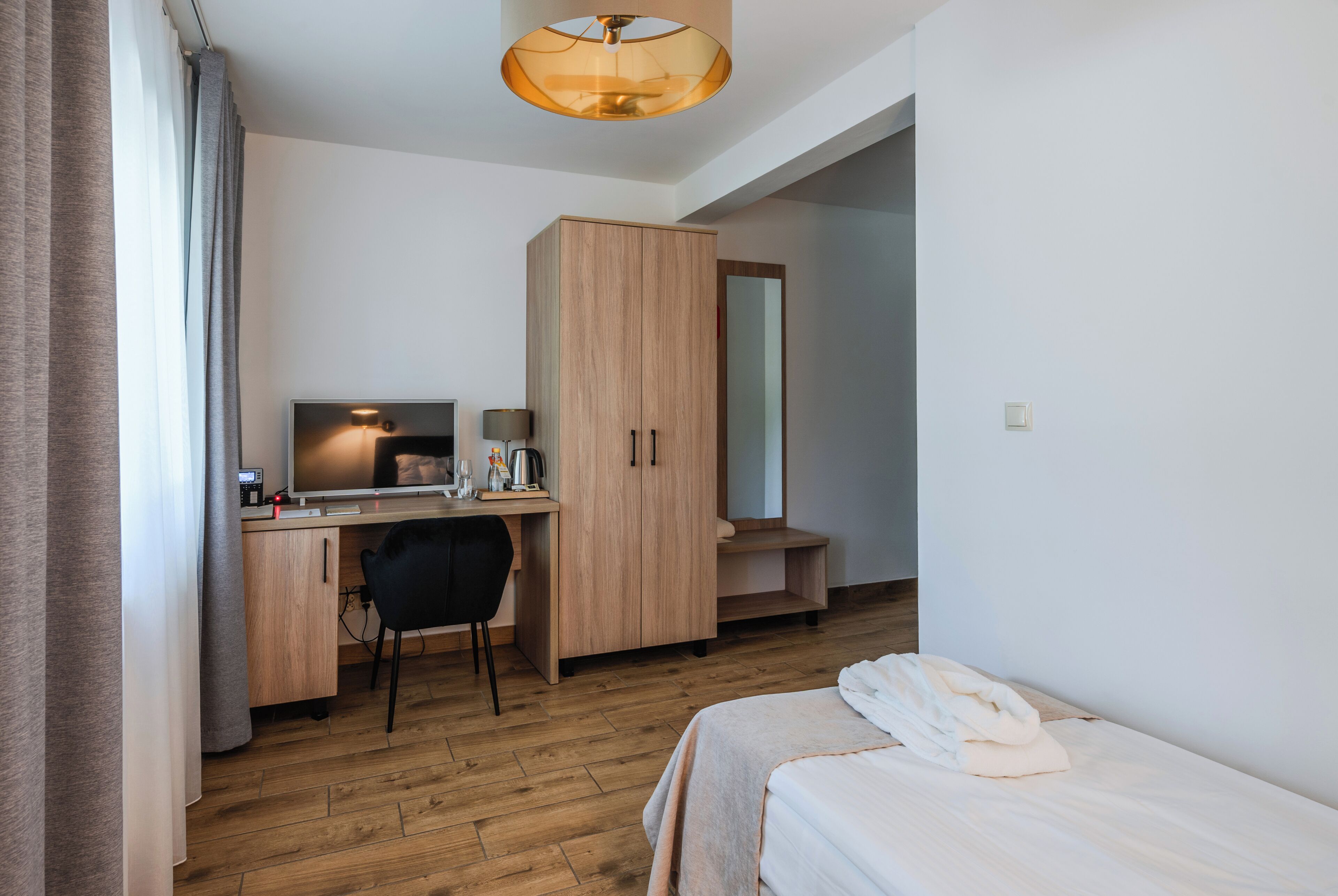 A hotel room with a wooden desk, a chair, a bed, a wardrobe, and a mirror.